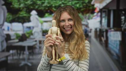 Woman holding a mannequin, smiling in a cafe with a blonde young model grasping a wooden artist puppet in an outdoor urban environment.