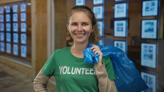 Woman holding a bag and smiling while participating in a cleanup event, blonde young volunteer carrying a plastic charity item in an indoor setting.