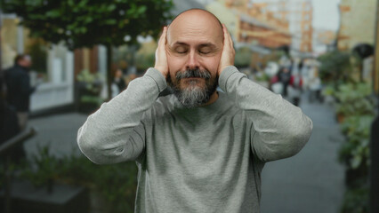Bald man with beard covers ears in urban street setting wearing gray sweater suggesting noise or...
