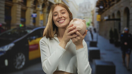 Young blonde woman smiling and kissing a piggy bank on urban street with taxi in background,...