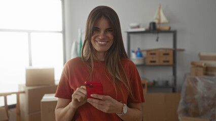 Woman using phone smiling in new home surrounded by moving boxes in bright living room showcasing a...