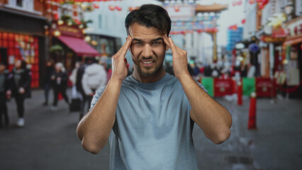 Young hispanic man feeling stressed in bustling city street with colorful decorations blurred in...