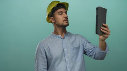 Young hispanic man wearing hardhat makes a video call holding a tablet against isolated green background, showcasing technology use in construction settings.