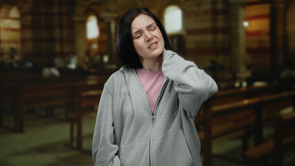 Woman experiencing neck pain while standing in a dimly lit catholic church interior with wooden...