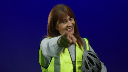 Woman smiling points forward wearing safety vest holds helmet against blue wall conveying...