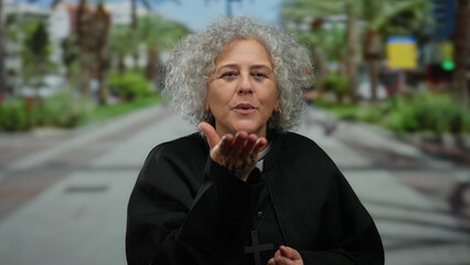 Elderly woman with grey hair and priest attire stands on an urban street, holding a cross,...