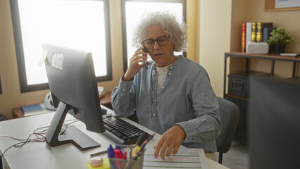 Woman in office talking on phone reviewing documents with focused expression at desk workspace.