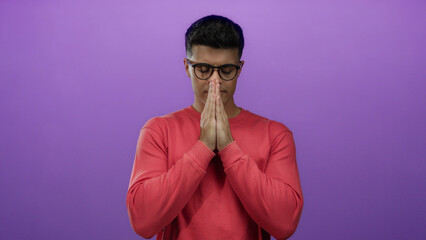 Young man with glasses in a red sweater praying against a purple background, hands together, in...