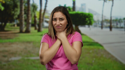 Young woman in pink shirt clasping hands to throat in a park with trees under a clear sky, showing...