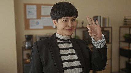 Woman smiling and making an okay gesture in an office setting surrounded by shelves and paperwork,...