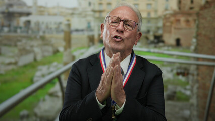 Senior man wearing suit and medal in roman ruins celebrating success, showing gratitude and...