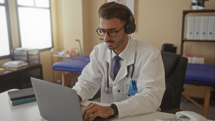 Man doctor wearing white coat stethoscope and headphones typing on laptop at clinic desk; focused...