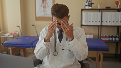 Young hispanic doctor wearing stethoscope at desk rubs eyes with palms in clinic; stress exhaustion...