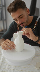 Man leans forward and holds brush to white vase in studio while steadying sculpted ceramic form;...