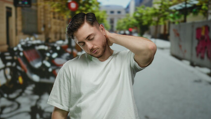 Young man with beard stands contemplatively on a lively city street, surrounded by vibrant greenery and urban scenery, reflecting modern outdoor life.