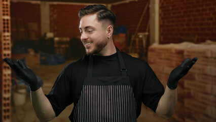 Young man with beard wearing apron and gloves smiling at construction site, suggesting indoor...