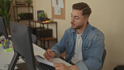 Young man with beard in office writing at desk beside computer focusing on work tasks in modern...