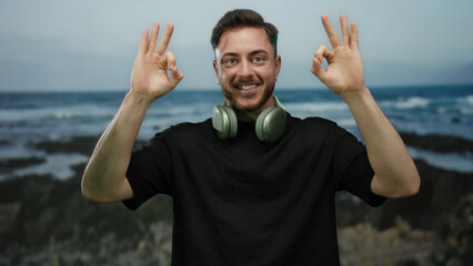Young man with beard and headphones gesturing ok sign at the seaside with a happy expression...