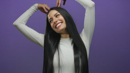 Naklejka premium Young hispanic woman forms a heart shape with her hands on a purple background, smiling joyfully in an isolated setting.