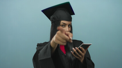 Graduated hispanic woman in cap and gown holds saudi passport against an isolated blue background,...
