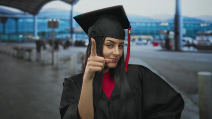 Young woman in graduation uniform gestures outdoors at an airport, embodying thoughtful success and...