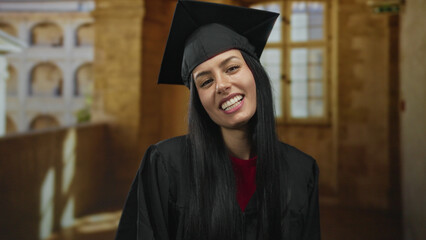 Young hispanic woman in graduation attire poses playfully indoors at an old university wearing a...