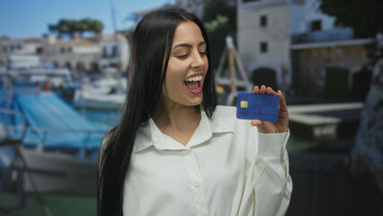 Woman smiling on a seaside port holding a credit card with boats and clear sky in the background outdoors.