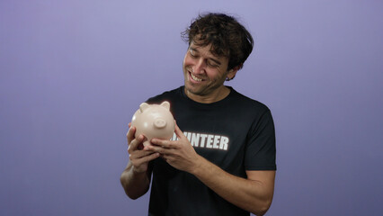 Hispanic man with a volunteer shirt smiling while holding a piggy bank against a purple background,...