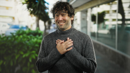 Young man standing outdoors in city street smiling with hands on heart showing gratitude and joy,...