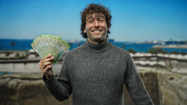 Hispanic man smiling on seaside beach holds russian rubles in the bright outdoors, suggesting financial freedom and travel joy.