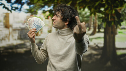 Hispanic man showing middle finger while holding polish zloty banknotes in a park outdoors,...