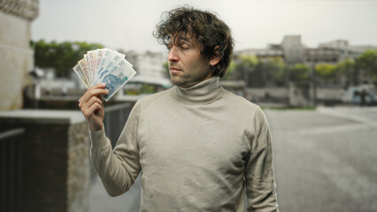 Hispanic man holding polish zloty banknotes on a city street outdoors, showcasing currency with urban backdrop, highlighting financial confidence in public space.