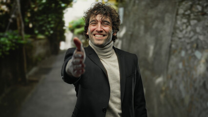 Fototapeta premium Young man smiling outdoors on city street extending hand for handshake in welcoming gesture, wearing black blazer and turtleneck sweater in casual urban environment.