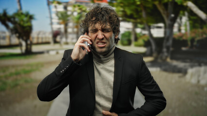 Young man outdoors in city holding phone with pained expression, wearing black jacket and beige turtleneck, suggests stomach ache amidst urban greenery backdrop.