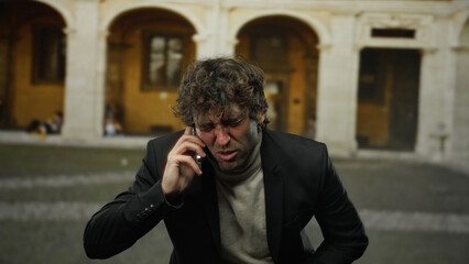 Young man with stomach pain talking on phone outdoors in city street, expressing discomfort while...