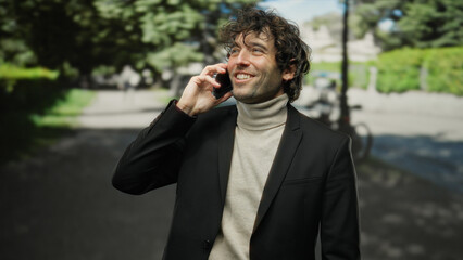 Handsome man talking on phone outdoors wearing casual jacket in sunny city setting, smiling brightly amidst urban landscape conveying a sense of joy and confidence