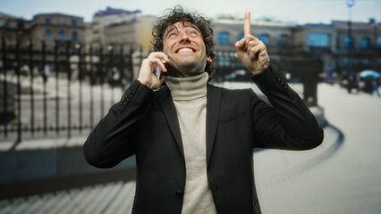 Man wearing a black suit smiling and talking on a phone while pointing upwards on a bustling city...