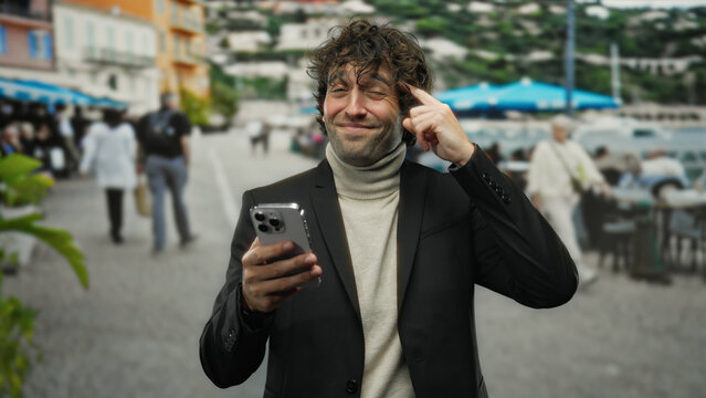 Young man outdoors touches head with finger while looking at smartphone in lively city street setting with people and colorful buildings in the background. - Powered by Adobe