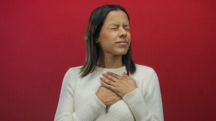 Hispanic woman in white sweater clutching heart in pain against isolated red background wall,...