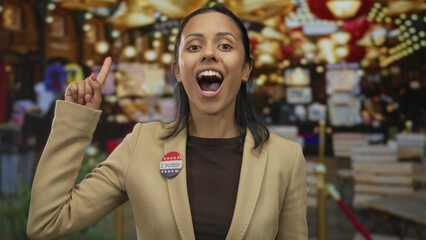 Hispanic woman making an idea gesture with an 'i voted' badge stands in an outdoor market with...