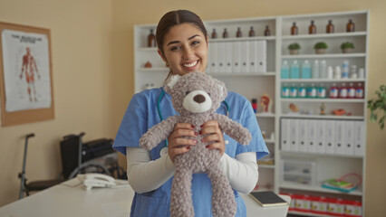 Hispanic woman doctor in clinic holding teddy bear smiling indoors with medical and care items...
