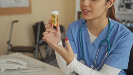 Hispanic woman doctor in hospital room holding medication bottle, wearing scrubs, examining pills...