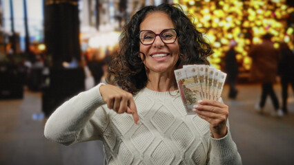 Woman outdoors holding russian banknotes and pointing down, surrounded by city lights on busy street, smiling in urban setting highlighting money and finance.