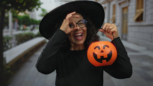 Middle age hispanic woman holds pumpkin basket and makes ok sign to eye in urban street; festivity celebration joy.