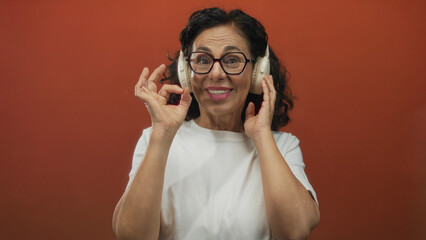 Middle aged hispanic woman wearing eyeglasses holds headphones and makes ok sign against red wall...