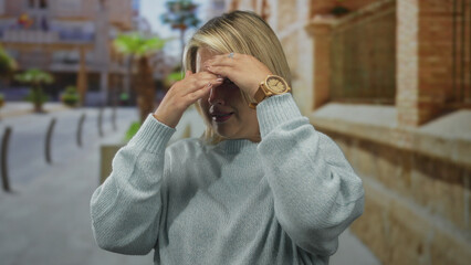 Woman covers face with hand on chest on a city street amid blurred storefronts and stone walls;...