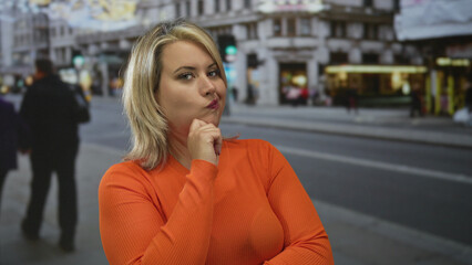 Blonde woman with hand on chin standing on a city street in front of building facades and...