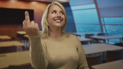 Smiling young blonde woman pointing finger toward camera in classroom with wooden desks and a large...