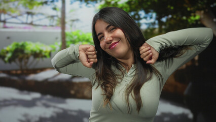 Smiling woman stretches arms upward in lush green park by a sunlit paved walkway under bright trees...