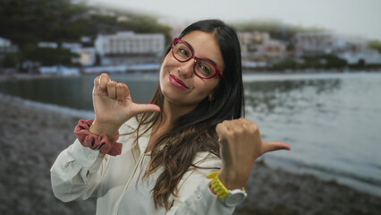 Hispanic woman wearing red glasses and a white blouse points her thumbs at herself on a sandy...